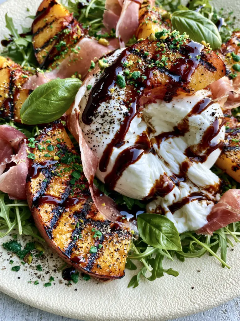 A high-angle shot of a Grilled peach salad with emerald-green pistachios, fresh basil leaves, and thin ribbons of prosciutto over a bed of wild arugula.