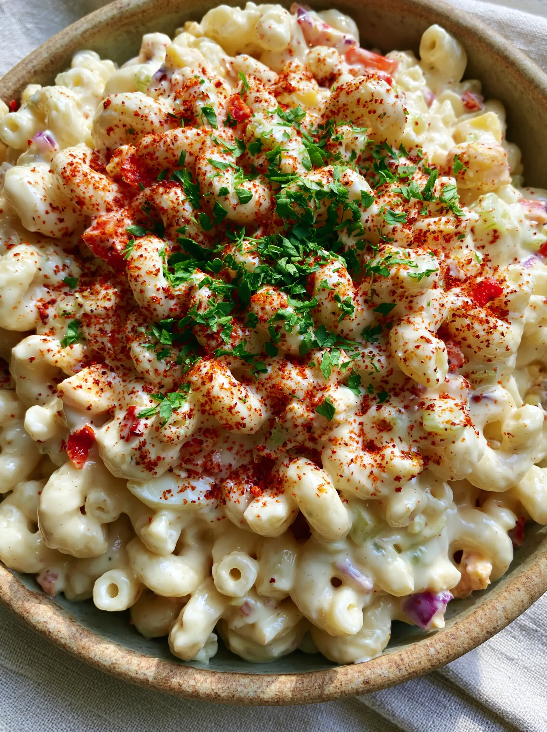 Top-down view of a rustic ceramic bowl filled with glossy homemade macaroni salad, garnished with parsley and paprika.