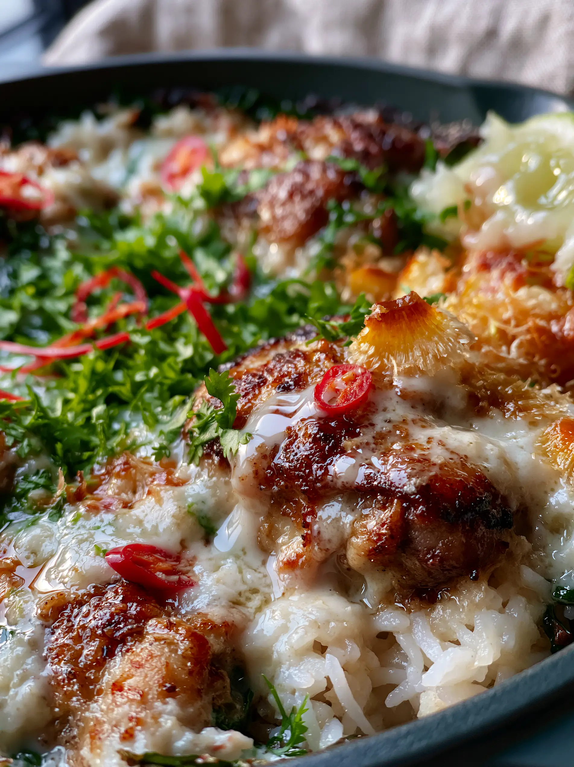 Extreme macro close-up of golden seared chicken chunks dripping in glossy coconut milk sauce with toasted flakes and red chilis. (Coconut Chicken Rice Bowl)
