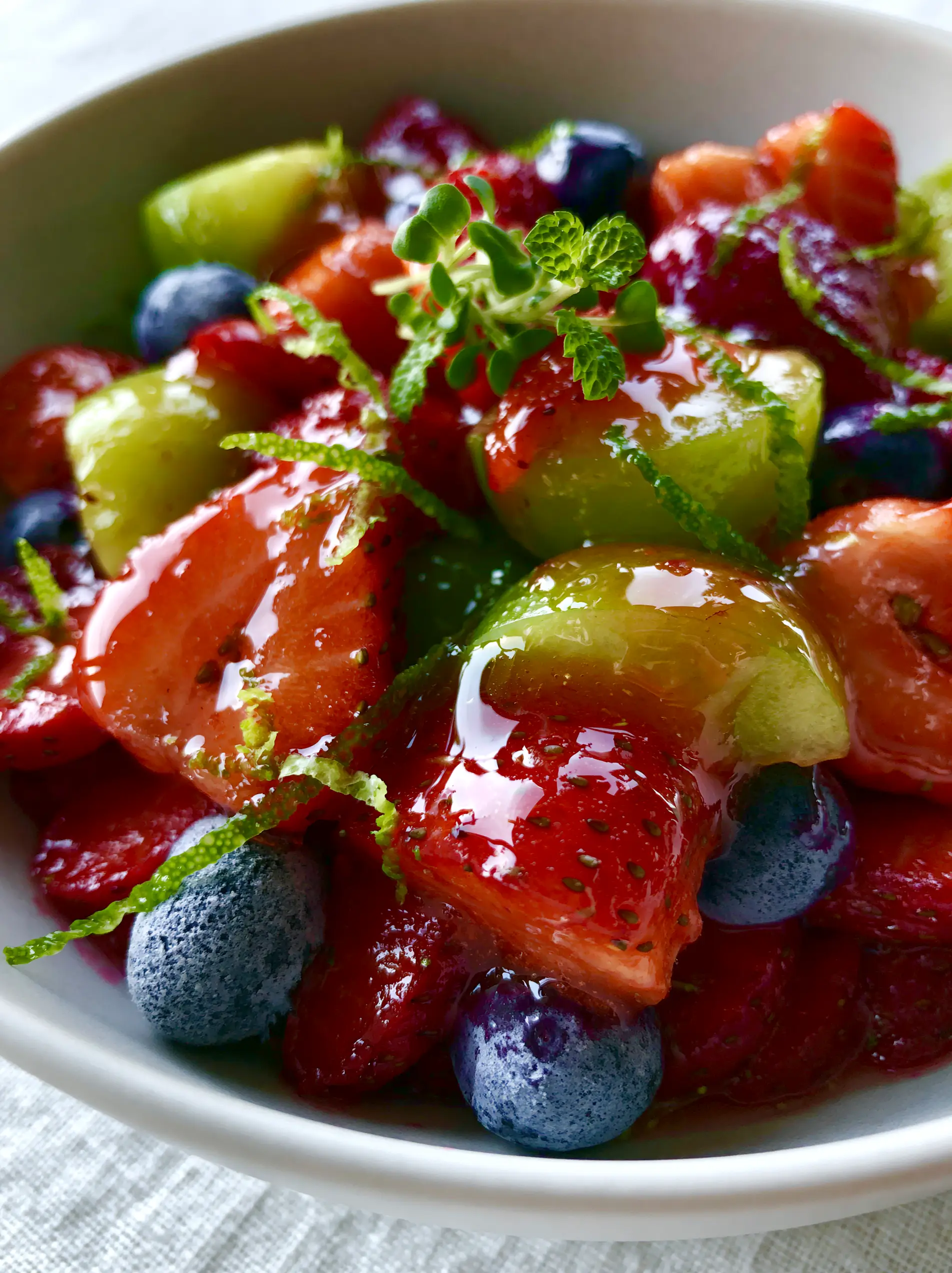 Extreme macro photography of honey lime fruit salad showing specular highlights on glistening strawberries and whole blueberries.